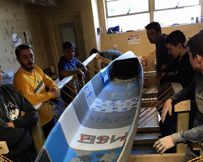 previous members standing around a canoe suspended by ropes that is themed as the different engineering majors at UConn